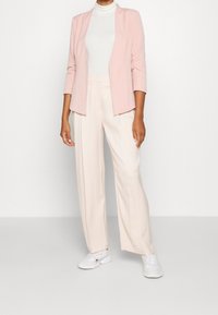 Blazer rose clair à manches trois-quarts, associé à un pantalon large crème et des baskets blanches, présentant une texture lisse et une coupe décontractée.