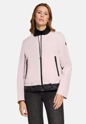 Frau trägt eine hellrosa Bomberjacke mit Reißverschluss, schwarzem gestepptem Saum und schwarzen Reißverschlusstaschen, kombiniert mit dunkler Jeans.