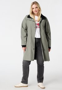 Femme portant un long manteau vert olive par-dessus un t-shirt à motif blanc, un jean noir et des baskets blanches, se tenant devant un fond uni.