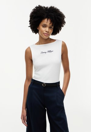 Kobieta o kręconych włosach, ubrana w białą, bezrękawnikową bluzkę z napisem „Tommy Hilfiger” oraz granatowe spodnie z wysokim stanem i czarnym plecionym paskiem.