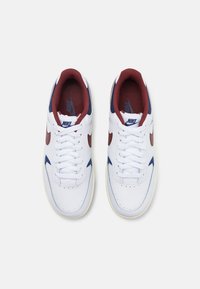 Biele tenisky Nike s marhuľovými a námorníckymi akcentmi, so hladkým koženým vrchom, okrúhlou špičkou a plochými šnúrkami. Textúrovaná stielka pre pohodlie.