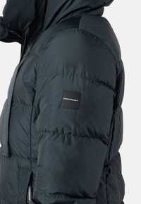 Puffet, mørkegrøn jakke med struktureret overflade, som har lynlås, hætte og et sort mærkepatch på ærmet.
