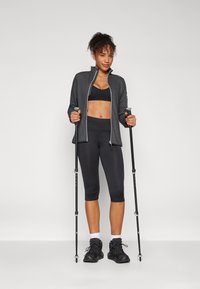 Giacca zip grigia scura sopra un reggiseno sportivo nero, abbinata a leggings neri lunghi fino al ginocchio e stivali da trekking neri, con in mano dei bastoncini da trekking.
