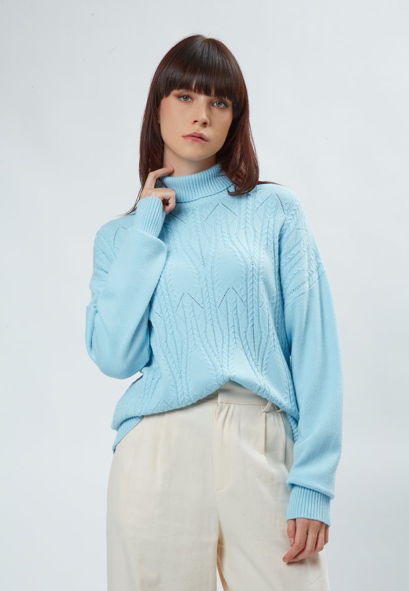 Maglione a collo alto lavorato a maglia di colore azzurro chiaro con un motivo testurizzato e polsini a coste, abbinato a pantaloni beige a vita alta.