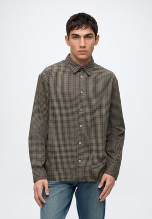 Junger Mann mit kurzem dunklem Haar, der ein braunes kariertes langärmliges Hemd und blaue Jeans trägt, steht vor einem schlichten weißen Hintergrund.