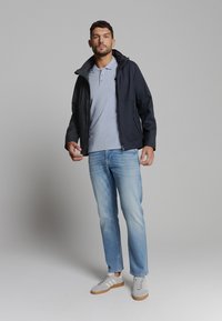 Marineblauwe windbreaker met capuchon, lichtblauwe polo, vervaagde blauwe jeans en grijze sneakers met witte strepen; comfortabele pasvorm, zachte textuur.