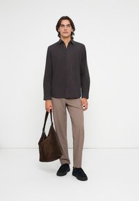 Barna kockás ing, bézs nadrág, fekete cipő és barna velúr válltáska. A modell egy fehér háttér előtt áll.