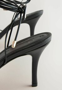 Escarpins à talons aiguilles noirs avec fines sangles à lacets et embouts métalliques argentés sur un fond neutre.