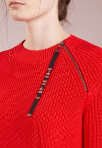 Pull rouge en maille avec un motif texturé, un col côtelé et un accent zippé accompagné d'une étiquette noire portant le nom de la marque.