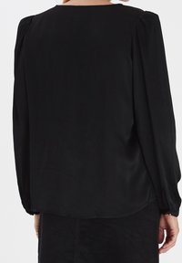 Černá blůza s dlouhým rukávem a kulatým výstřihem, nařasenými ramenními švy a zakřiveným spodním lemem. Hladká textilie s minimálním designem.