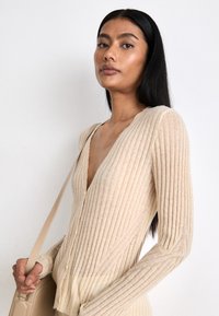 Cardigan beige a costine con scollatura a V profonda, maniche lunghe e chiusura a bottoni. Tessuto morbido con strisce verticali, abbinato a una borsa beige.
