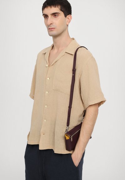 Beige short-sleeve shirt in linen with pocket, paired with a small maroon leather crossbody bag featuring a yellow interior and adjustable strap.