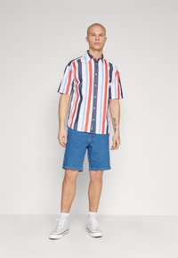 Camisa de manga corta con botones y rayas horizontales en rojo, azul marino y blanco; combinación con pantalones cortos de mezclilla azul y zapatillas de color claro.