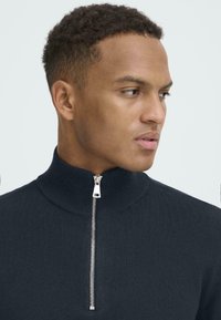 Tamnoplavi zip-up pulover izrađen od rebrastog materijala, s visokim ovratnikom i srebrnim zatvaračem. Glatka tekstura i uski kroj.