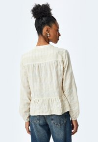 Blusa a righe color beige chiaro con orlo peplum, maniche lunghe e vestibilità rilassata. Realizzata in tessuto testurizzato con una leggera lucentezza.