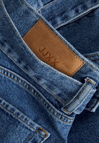 Denim jeans featuring a light to medium wash, a brown leather patch embossed with "JUXX," and visible contrasting stitching.
