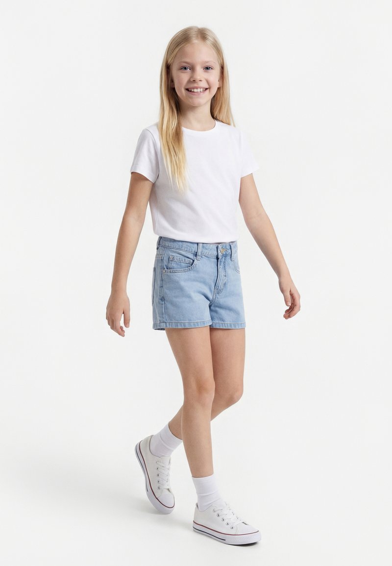 Usmievavá dievčina s dlhými blond vlasmi, ktorá kráča, oblečená v bielom tričku, svetlomodrých džínsových šortkách, bielych členkových ponožkách a bielych teniskách.