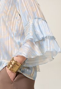 Blusa azzurro chiaro, trasparente, con maniche a volant e motivo a righe arricchita da dettagli dorati. Accompagnata da un largo bracciale in oro.