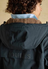 Čierna nepremokavá bunda s golierom lemovaným kožušinou. Má modro-biely pruhovaný vnútorný golier. Na zadnej strane je potlačený text „JOULES“.