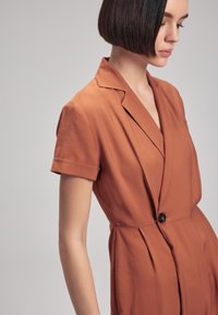 Vestido color óxido con mangas cortas, un cuello con muesca y un cierre de un solo botón. Presenta pliegues y una textura de tela suave.