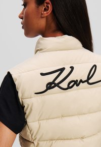 Beige gesteppte Weste mit hohem Kragen, auf der Rückseite mit schwarz gesticktem "Karl". Der Stoff hat eine glatte Textur und ein gesteppte Design.