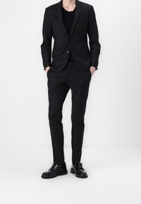 Svart skräddarsydd kostym med en enkelknappad jacka och slim-fit byxor. Bärs över en svart rundhalsad tröja, matchat med svarta skor.