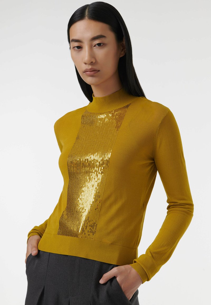 Senfgelber Pullover mit Mockneck und einem mit Pailletten verzierten Mittelteil. Lange Ärmel und eine taillierte Silhouette. Die Textur ist glatt.