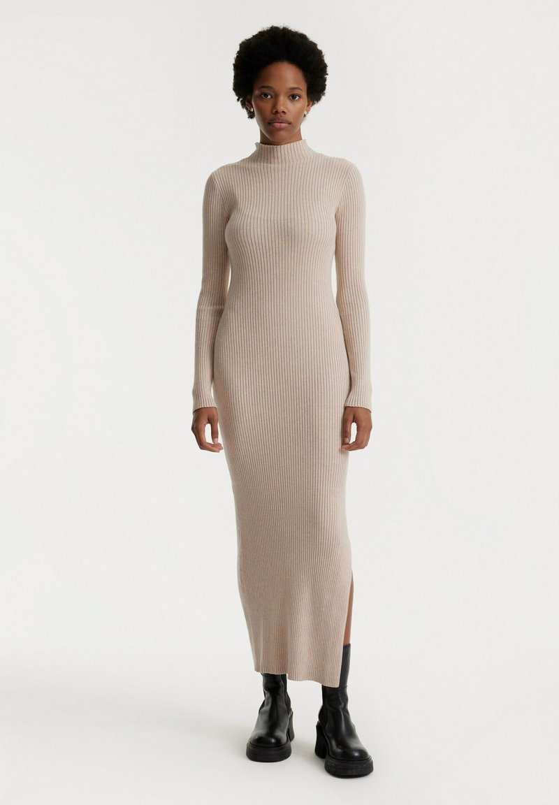 Lange geribbelde jurk in lichtbeige, met een hoge hals, lange mouwen, splitten aan de zijkanten en een getailleerd silhouet. Gekocht met zwarte chunky laarzen.
