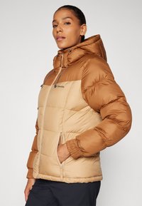 Veste puffer marron avec capuche, dotée d'un design bicolore. Comprend une poche zippée, des poignets élastiques et un logo sur la poitrine.
