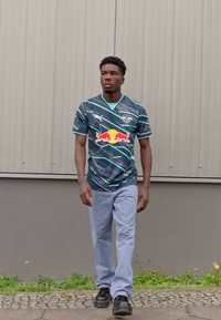 Fekete sportmez türkiz és fehér ferdén futó vonalakkal, Red Bull logóval, világoskék nadrággal és fekete cipővel párosítva. Világosszürke háttér.