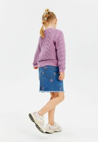 Lila stickad tröja med ett diamantmönster, kombinerad med en blå denimkjol som har regnbågsbroderi. Beige sneakers med en texturerad sula.