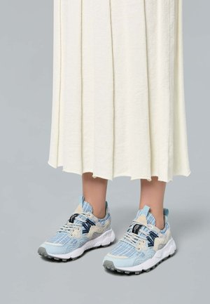 Osoba nosząca jasnoniebiesko-beżowe dzianinowe tenisówki z białymi podeszwami i sznurowadłami, w zestawieniu z kremową plisowaną spódnicą midi na szarym tle.