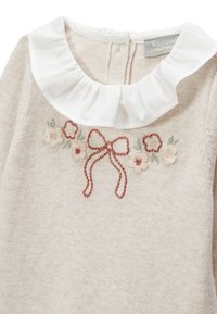 Pull beige clair avec un col à volants blanc et une broderie florale en rose et vert. Comprend un motif de nœud brodé au niveau de l'encolure.