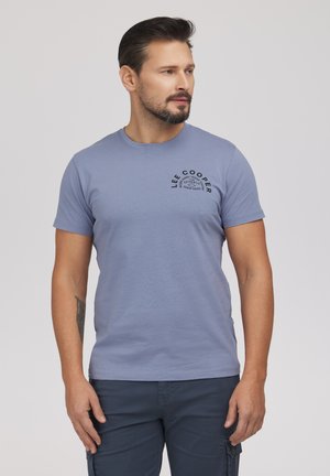 Mężczyzna z brodą i tatuażem na przedramieniu, ubrany w jasnoniebieską koszulkę Lee Cooper i ciemne spodnie, patrzący w bok na jednolitym tle.