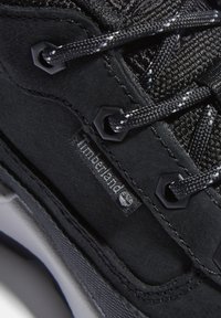 Zapato deportivo negro con una parte superior de ante texturizado, detalles de malla, ojales hexagonales y un logotipo de Timberland visible en el lateral.