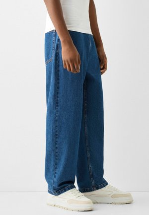 Person trägt lockere blaue Jeans, weiße Turnschuhe und ein weißes Hemd, steht vor einem schlichten hellen Hintergrund.