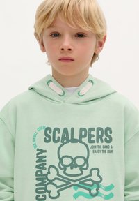Felpa verde chiaro con grafica di teschio e ossa incrociate, cappuccio regolabile con coulisse e polsini a costine. Realizzata in morbido tessuto di cotone.
