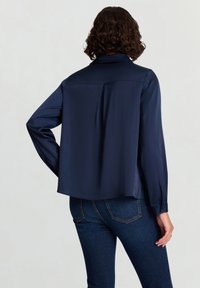 Chemise en satin bleu marine à manches longues, col pointu et détail de pli dans le dos. Texture lisse, coupe légèrement ample, et poignets à boutons.