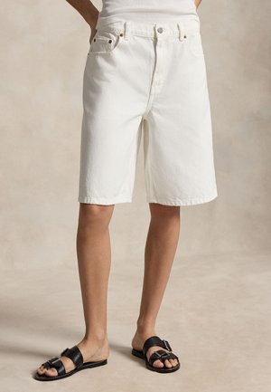 Asmuo dėvi baltus džinsinius šortus iki kelių ir juodus atvirus sandalus su sagtimi prieš neutralų foną.