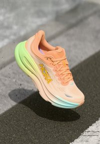 HOKA løpesko med åpen mesh-overdel i ferskenfarge med gule detaljer, dempet såle med gradient fra grønt til lyseblått, og intrikat tekstur.