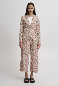 Blazer à motif floral et pantalon large dans des couleurs pastel, avec des teintes de rose, vert et beige, en tissu doux et texturé. Sandales plates.