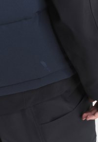 Mornarica jakna s teksturiranim materijalom, koja ima glatki dio ispod i suptilnu oznaku detalja. Vidljive džepove dodaju funkcionalnost.