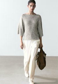 Woman walking forward wearing a loose gray knit sweater, cream wide-leg pants, white shoes, holding a large brown bag with a patterned scarf.