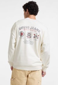 Krém színű pulóver, melyen a hátulján a "GUESS JEANS AMERICAN TRADITION" felirat és logók találhatók. Lazább szabású, bordás mandzsettagal.