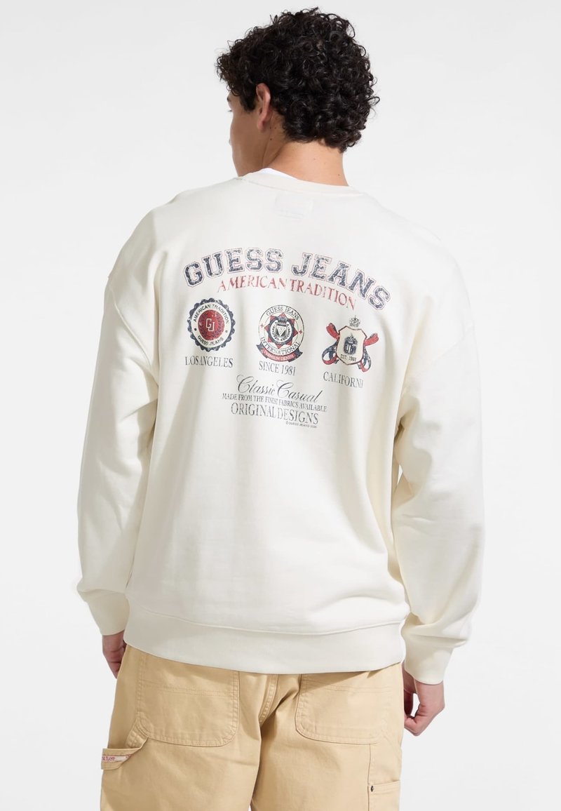 Krém színű pulóver, melyen a hátulján a "GUESS JEANS AMERICAN TRADITION" felirat és logók találhatók. Lazább szabású, bordás mandzsettagal.