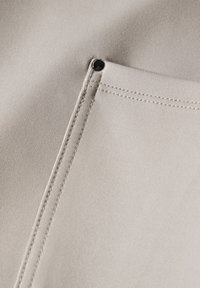 Beige tyg med en slät yta, med en sydd söm och en metallögla nära en liten ficka.