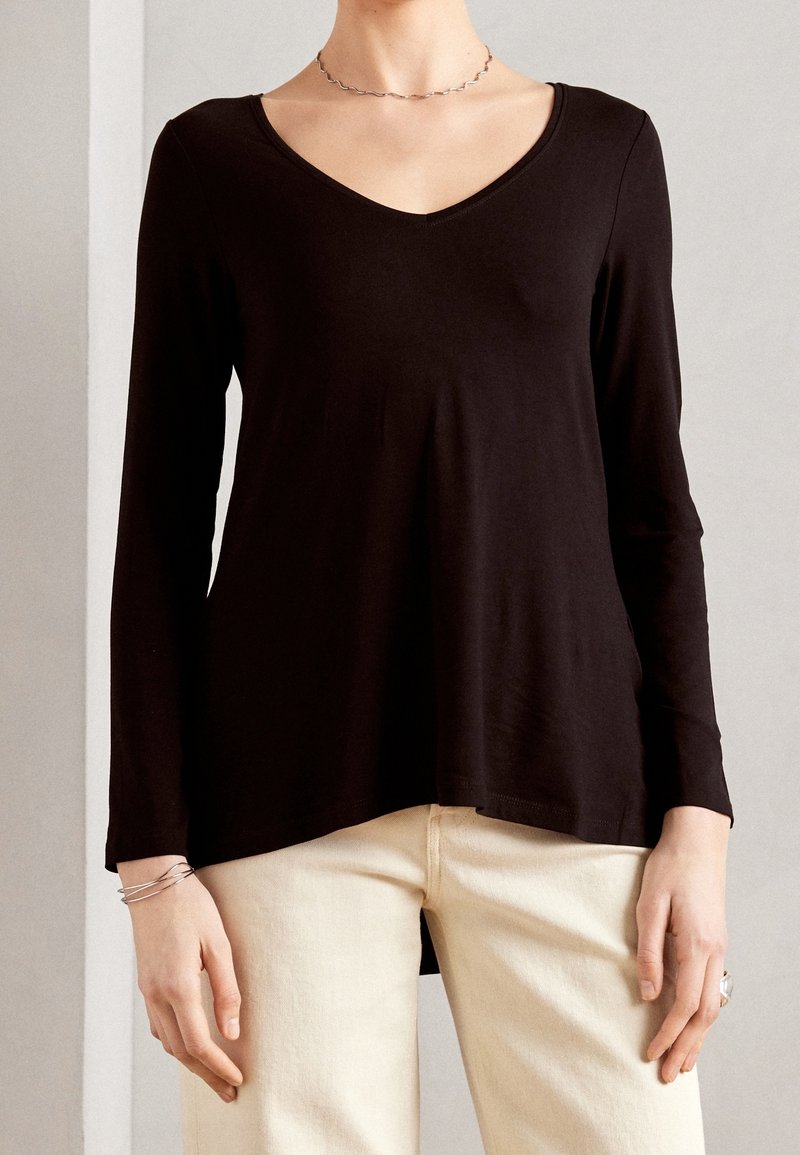 Top negro de manga larga con escote en V, hecho de tela suave. Presenta un ajuste relajado y dobladillo curvado, combinado con pantalones de color crema.