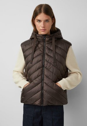 Braune gepolsterte Weste mit Kapuze, versehen mit einem Zickzack-Steppmuster und einem Reißverschluss. Cremefarbene strukturierte Ärmel, kombiniert mit dunklen Jeans.