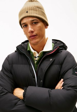 Junger Mann mit beiger Strickmütze und schwarzer Steppjacke, die Arme verschränkt, gekleidet für kaltes Wetter.