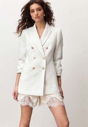 Kvinde med bølget hår iført en hvid dobbeltradet blazer og hvide shorts med blonder, der står mod en ensfarvet baggrund.